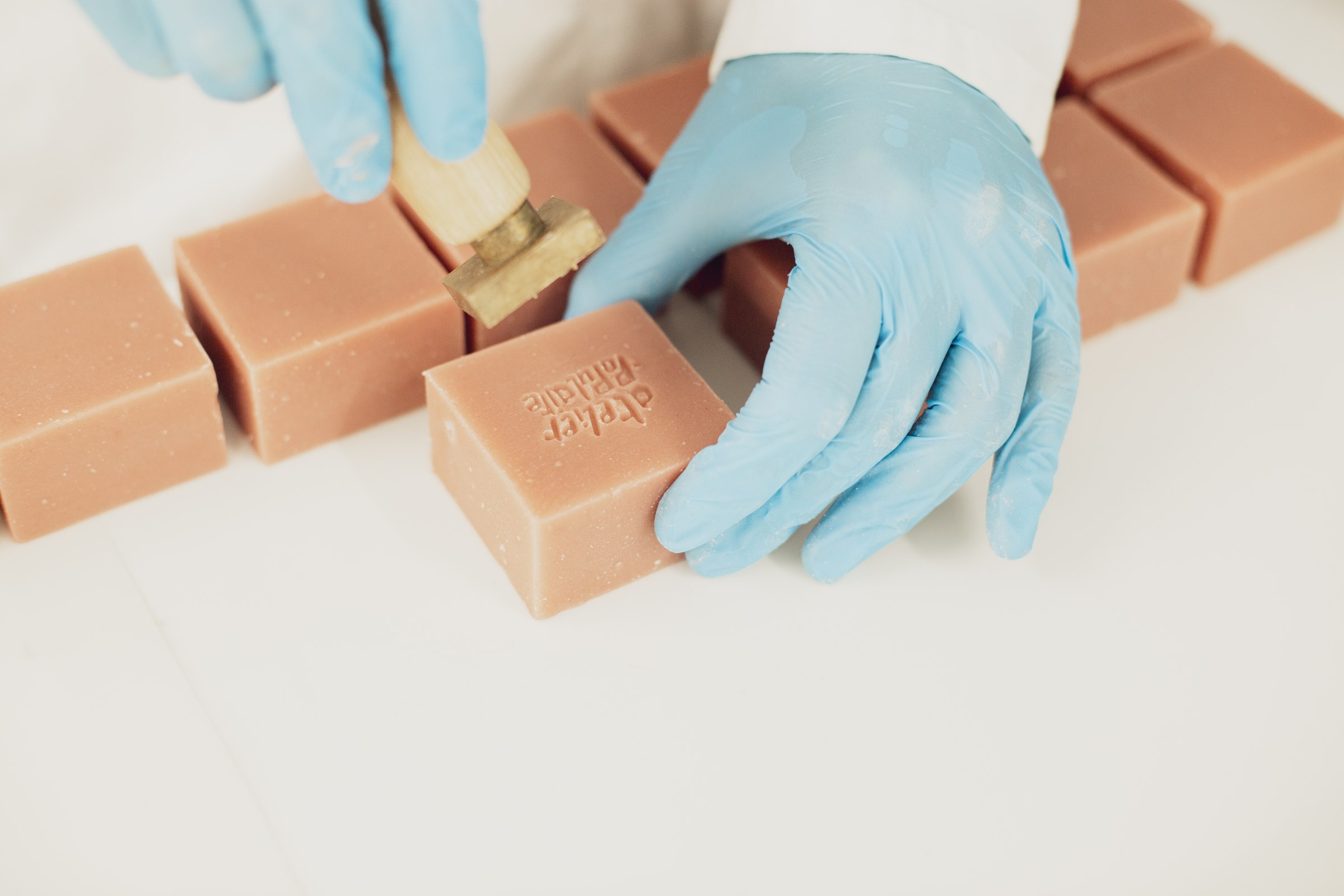 Des mains gantées tamponnent un savon, parmi les pavés alignés sur le plan de travail