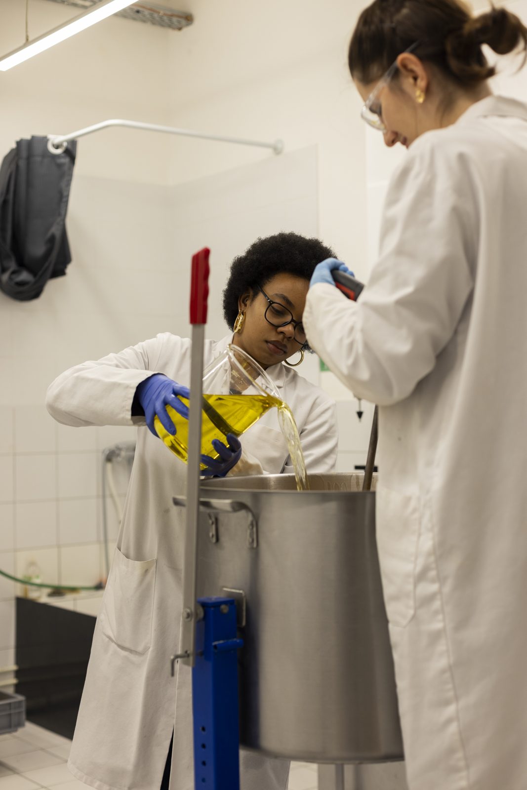 Two workshop staff in lab coats mix ingredients in a production vessel